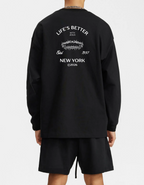 A person shown from the back wearing a black long-sleeve sweatshirt with a white graphic print on the back reading 'LIFE'S BETTER NEW YORK' and black shorts against a light gray studio background.