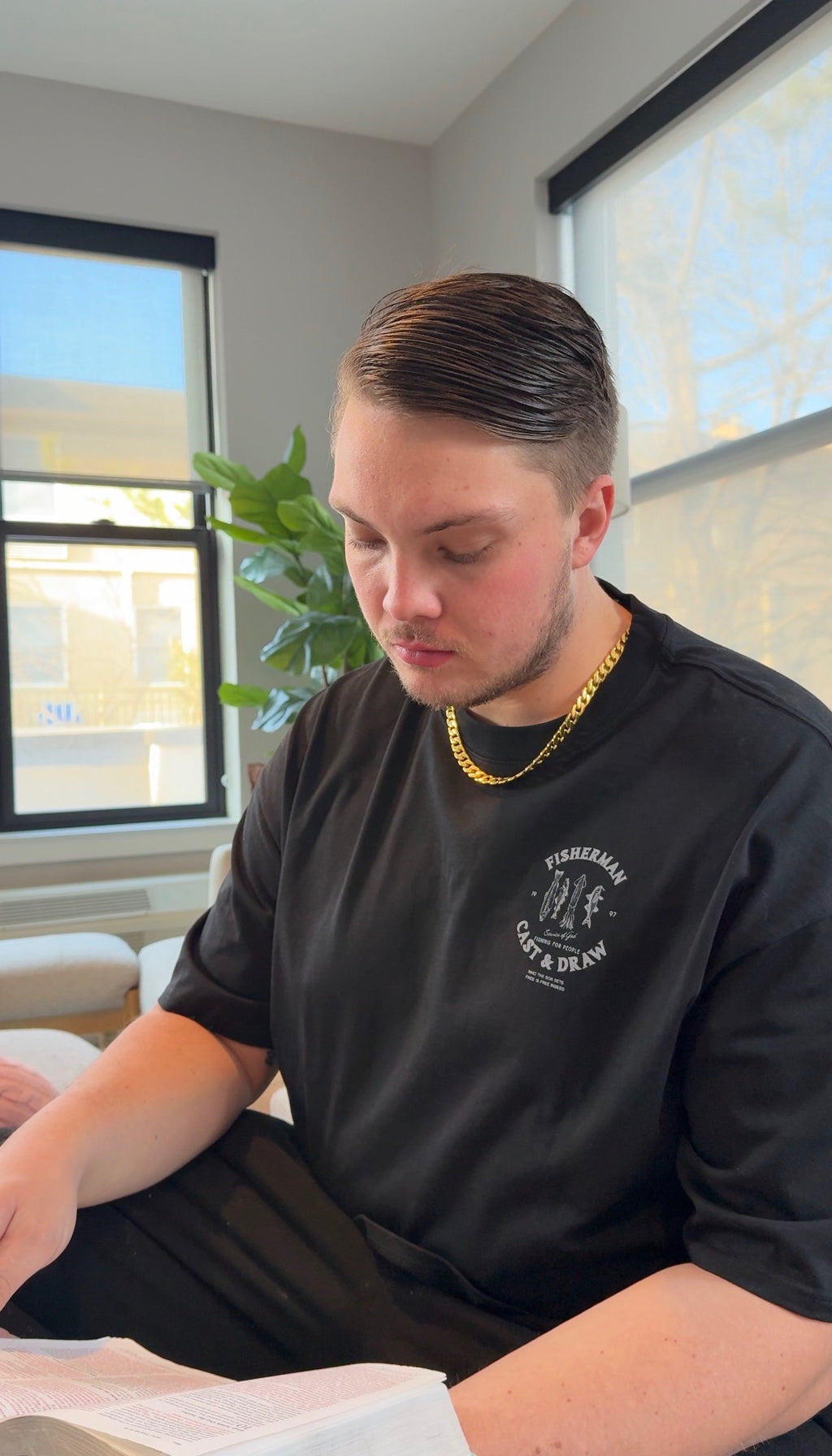 Man wearing a black t-shirt with a logo, sitting indoors near a window.