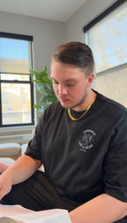 Man wearing a black t-shirt with a logo, sitting indoors near a window.
