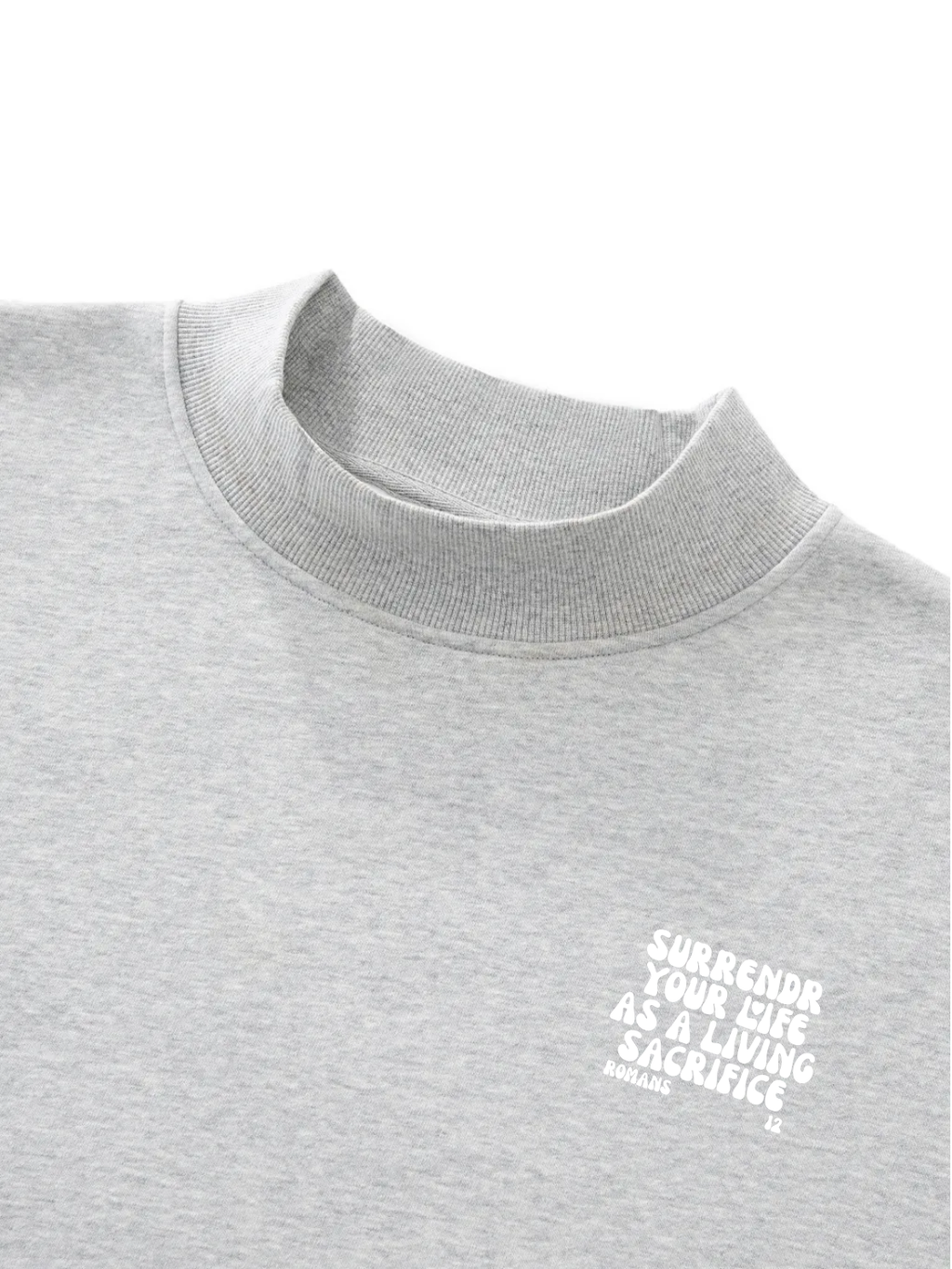 Close-up of a light heather gray crewneck sweatshirt with a small white graphic text on the left chest reading 'SURRENDER YOUR LIFE AS A LIVING SACRIFICE' and 'ROMANS 12' below it.