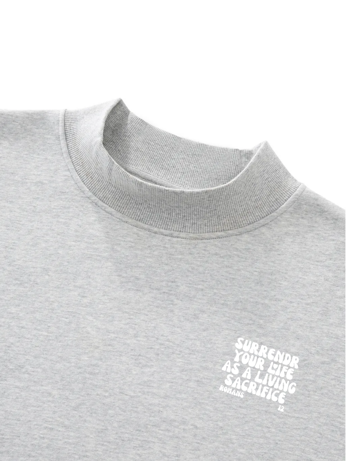 Close-up of a light heather gray crewneck sweatshirt with a small white graphic text on the left chest reading 'SURRENDER YOUR LIFE AS A LIVING SACRIFICE' and 'ROMANS 12' below it.